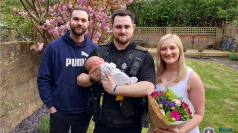 PC Ronnie Barrett holds a month old baby in the middle of her parents. They are all in a garden
