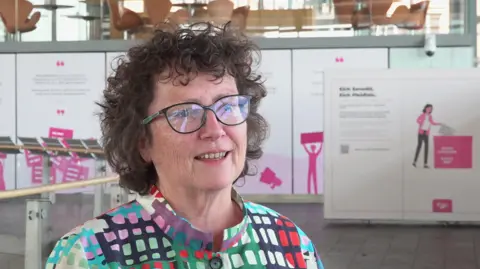 Elin Jones wearing glasses and a colourful patterned jacket is photographed inside the Senedd building.
Pink election-themed displays and ballot box graphics can be seen in the background.