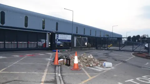CPS Two large orange traffic cones stand next to a pile of bricks and two black fire extinguishers, in front of a large grey factory building.