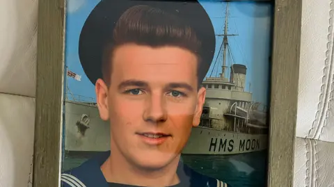 Wally Newman Wally Newman aged around 17 in Naval uniform, behind him is a grey war ship, MHS Moon, the picture has been colourised. Newman has brown hair swept back over his head and a blue Navel cap on. 