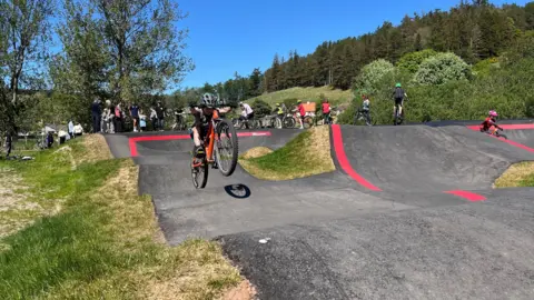 A child rides a bicycle on the pump track in the background with several other cyclists