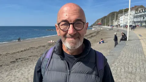 A man in dark clothing wearing spectacles and a backpack standing in front of the sea.