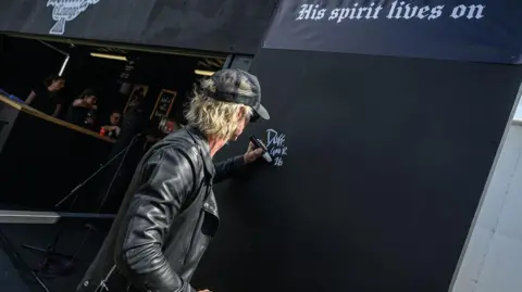 Katja Ogrin/Getty Images A man with his back to the camera, signing "Duff, GNR, 26" on a wall with a white pen. He is wearing a black leather jacket and a black baseball cap.