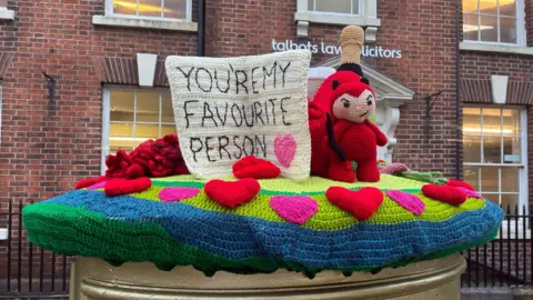 A knitted representation of a red devil and a Valentines card. There is a brick building in the background. They are sat on top of a gold post box.