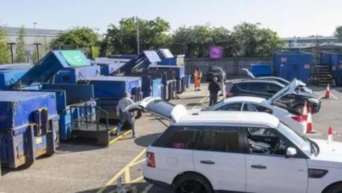 North Yorkshire Council A group of cars parked at a household waste recycling centre with a number of large waste containers in blue to the left of the image.