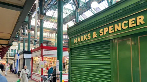 An indoor market scene featuring a green Marks & Spencer stall with its shutter closed in the foreground. The market has a high glass roof supported by ornate green metal columns. In the background, other stalls are open and decorated, including one with red trim displaying various goods.