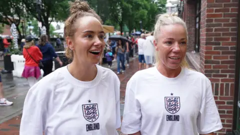 Two women wearing England football shirts smiling together on a Boston street 