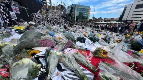 Andy Greenwood/BBC Bunches of flowers, including white roses and sunflowers lying outside St James' Park in Newcastle, together with club scarfs and T-shirts. People are looking at the tributes.