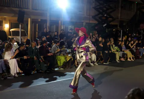 Hollywood 2025 at Paramount Studios in a gold suit with flaired trousers, purple trim and fushia pink blouse and socks. The audience claps in the background