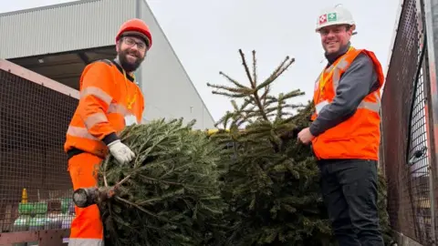 Dougie Mac Two men in orange high-vis tops and bottoms and hard hats smile as they hold Christmas trees on the back of a lorry.