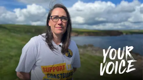 A woman with long, brown hair and glasses in a white T-Shirt which says "Support Your Coastguard". She is standing in front of a blurred coastline with clouds in a blue sky above her. There is a Your Voice logo on the image.