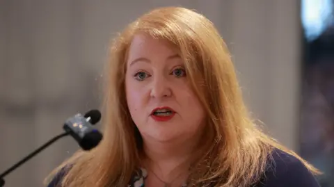 A long red-haired woman speaks into a black microphone, wearing red lipstick. She wears a purple cardigan with a white and teal shirt underneath. 