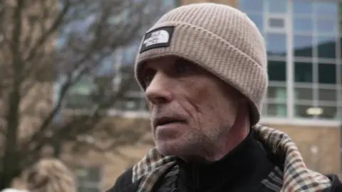 A man wearing a brown hat and jacket speaks to a reporter who is not on camera