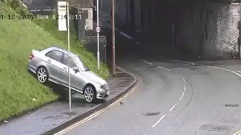 Image take from a CCTV video shows a silver car bouncing onto a stretch of pavement heading towards a main road from a grass hill.