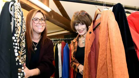 Quay Theatre, Sudbury Krystle and Julie are inside the costume store standing behind a number of garments hanging on a rail. Krystle is on the left wearing a black top and Julie is on the right wearing a black top and red and black checked jacket over the top.