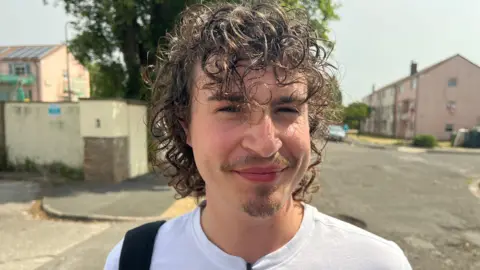 Archie Byron is smiling at the camera. the image is a head and shoulders shot of Archie. He has dark curly hair and a small beard on his chin. He is wearing a white T-shirt and has a bag over one shoulder. He is stood in an urban area with a block of flats in the background. 