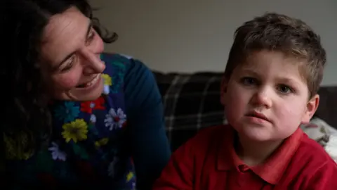 A five year old-boy with blue eyes and short brown hair looks into camera, while wearing a red polo shirt. His mum, wearing a blue dress with bright red and yellow flowers on, looks at her son adoringly while sat next to him on him on a sofa.