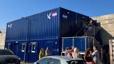 Jon Wright/BBC A block of four blue shipping containers, with people standing outside.