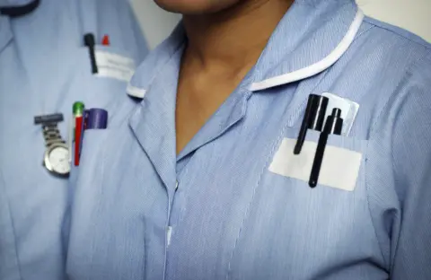 A stock image of two nurse's torsos both wearing blue uniforms with pens in breast pockets and a fob watch
