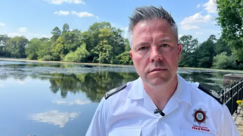 BBC David Lynch from Lincolnshire Fire & Rescue stands next to a lake surrounded by trees. He has greying hair and is wearing a white shirt with badge embroidered on the front.