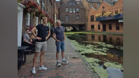 Supplied Two men stand on a brick pathway next to a canal lined with brick buildings. 