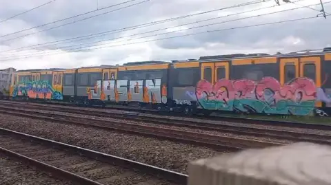 Three metro carriages on a rail sit with graffitti on each, the artwork stretches from the bottom edge of the carriage up to part way through the windows of the trains. The middle carriage has the name Jason painted in silver with fire around the edges while the other two have illegible words coloured in pinks, greens, blues and oranges. 