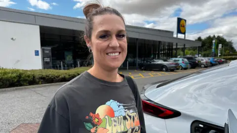 A smiling woman with her hair in a bun stands beside a white electric vehicle connected to a charging cable at Lidl's Barton-upon-Humber store. The supermarket building and Lidl sign are visible in the background, with several parked cars in the car park under a cloudy sky. Nikki is wearing a retro-style colourful T -shirt.