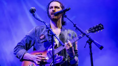 Hozier performing onstage with his guitar. He is wearing a denim jacket.