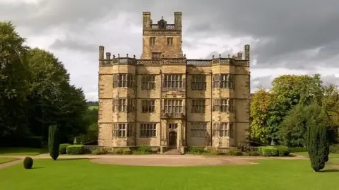 The imposing three-storey stone-fronted Gawthorpe Hall, with a tower in its centre on an additional level. The hall is surrounded by a neatly mown lawn, trees and shrubs.