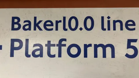A close-up of a wall sign reading “Bakerl0.0 line Platform 5”, with the zero in “0.0” in place of two letter o's in Bakerloo.