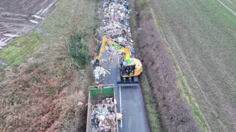 A huge pile of waste in the background is being handled by a yellow digger which is scooping the waste into a large green container. The pile of waste visibly blocks the road.