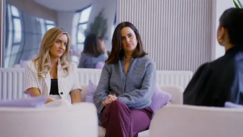 IWC MEDIA/BBC SCOTLAND Agency co-founders Amy Moore and Kirsten Cameron sit together on a fashionable cream sofa with purple boucle cushions with a mirror behind them. They are talking to an influencer who we can't quite see - just the back of their head in the foreground. Amy is wearing a white trouser suit with black wide pinstripes and Kirsten is in purple trousers and a lilac/grey cardigan.