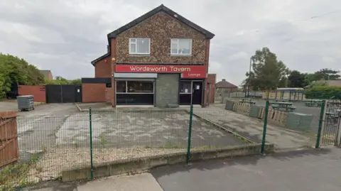 A building with a red sign across it, which reads Wordsworth Tavern. Above the black front door is a sign which says Lounge. There is a concreted area in front of the building and a green wire fence. To the right there are picnic tables and a gate.