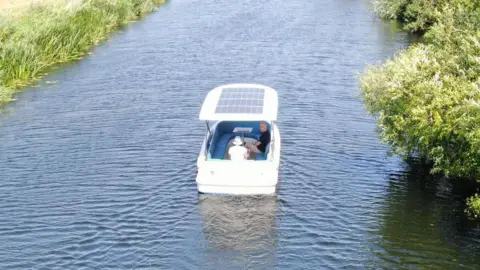Torkmar Tim Knox and his wife Sue travelling on the boat. It is a small white vessel with solar paneling on its roof.