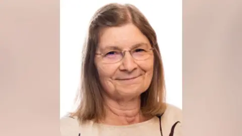 A woman with long light brown hair and wearing glasses looking into the camera. She is wearing a light brown jumper.