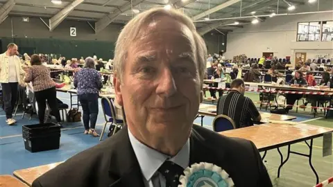 Reform's Robert Parsonage. He is wearing a black suit, blue shirt and has a Reform rosette on his chest. He has short grey hair.