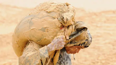 Reuters A man is bent over with a sack on this back. His clothes are covered and hands are covered in mud. In the background is a blurred dirt landscape.