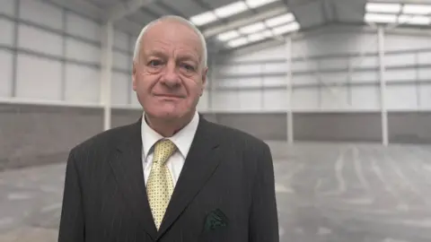 LDRS Milan has short grey hair and is wearing a black suit and yellow tie. He is in an industrial unit. 