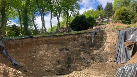 A large excavated earthen pit along the banks of a garden. Trees can be seen along the intact section of bank and a home can be seen in the background.