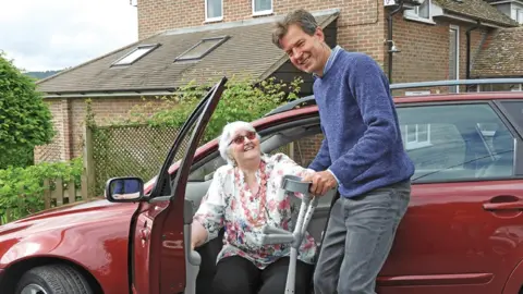 A man in a blue jumper and grey trousers is helping an older lady out of a red car. He is holding crutches for her and smiling to camera. There is a brick house in the background 