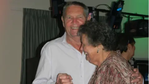 Family handout Bob and Sylvia dance together at a function. Bob is wearing a white shirt and Sylvia is wearing a multi-coloured top.