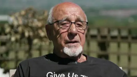 Head and shoulders image of John Morris sitting in the garden in the sunshine in a black tshirt. He is wearing glasses and has short white receeding hair and a goatee