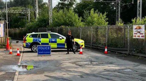 Someone is standing next to a police car, which is side on to the camera. Cones and a sign saying 'police road closed' are in front of the car. There is a high metal railing to the right of the image with greenery and train lines behind it.