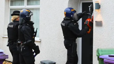 A police officer wearing a protection vest and helmet is using a saw to break down the front door of a house. Two police officers are standing behind them. 
