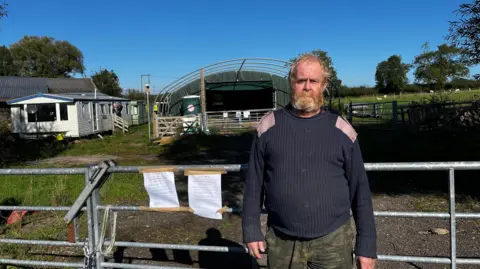 A man wearing a knitted jumper and combat trousers stands in front of a five-bar gate with two eviction notices on it. In the background is an agricultural shed and two mobile homes. 