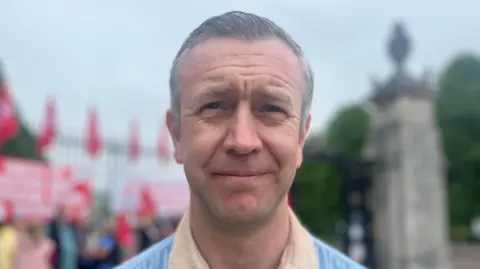 Kieran Ellison who has short grey/brown hair and a blue shirt with a tan collar he is smiling at the camera in front of a blurred background of a gate with people holding signs 