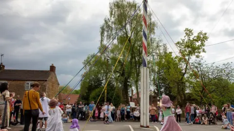 Electric Egg A tall maypole which is stripy blue, white and red has coloured ribbon coming out of it which children are pulling and dressed in traditional clothing. People can be seen stood around the maypole watching the children.
