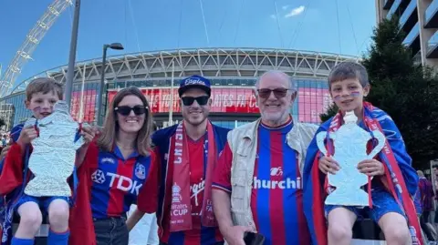 Robin Wicks A family of five Crystal Palace fans wearing club shirts pose outside Wembley Stadium, holding replica tinfoil trophies, with the stadium in the background.
