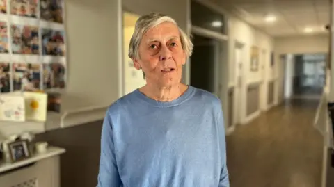 A woman with grey hair in a blue jumper is looking at the camera. The corridor is blurred behind her. On the wall are several cards and photos.