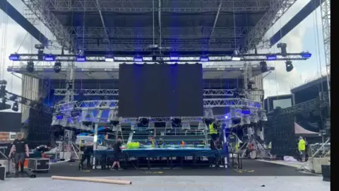 Dino Cabras Workers in hi-vis tops around the ring area which has been placed in the centre of the Portman Road pitch. There is a large black screen hanging from a large metal gantry.